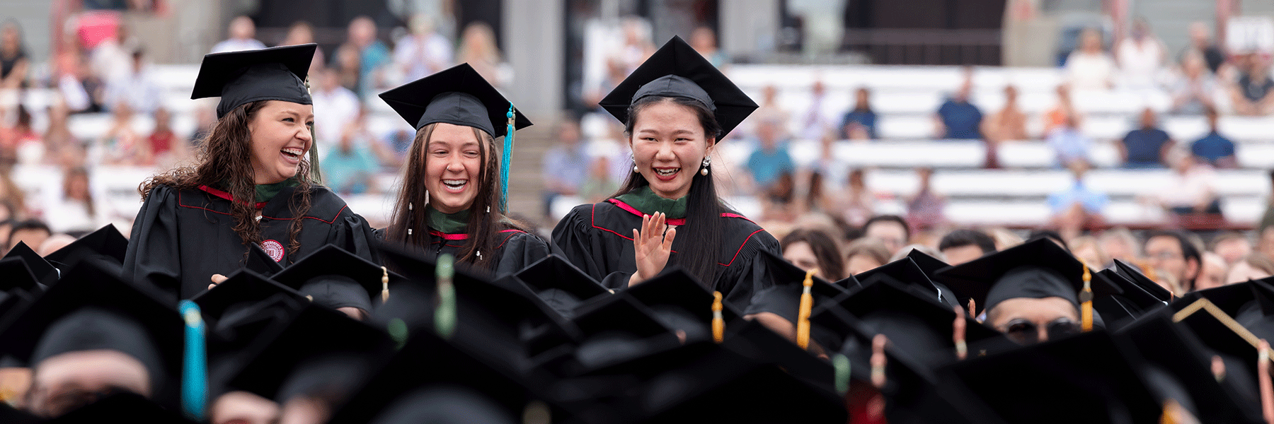 Three female graduates smiling as they stand up in a crowd at commencement.