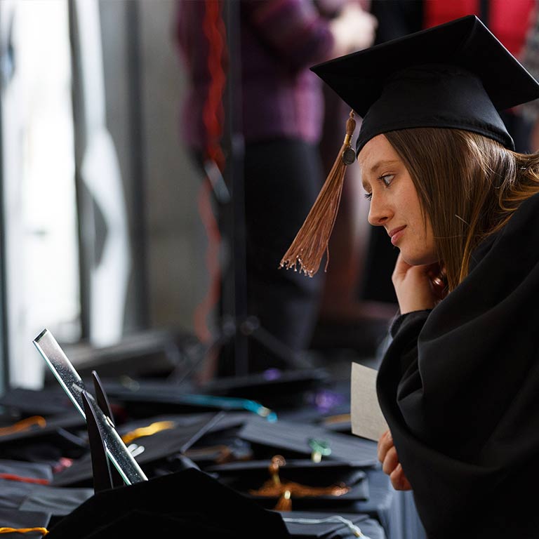 A student trying on a cap and gown at GradFair looks into a mirror set on a table.