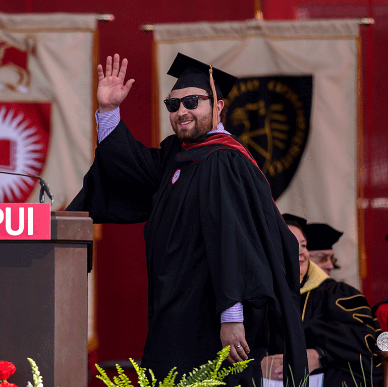 A male graduate in master's commencement attire smiles and waves from the stage.