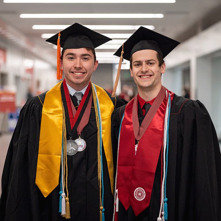 Two male Purdue and IU graduates pose together and smile wearing their graduation robes, stoles, and honor cords.