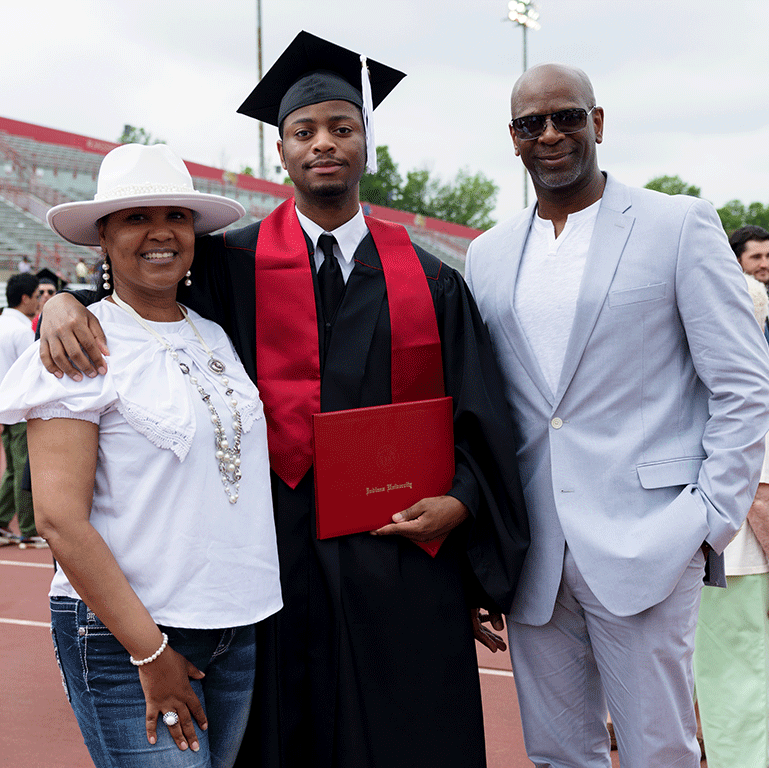 A male graduate smiles between his parents at the Commencement ceremony, holding a diploma