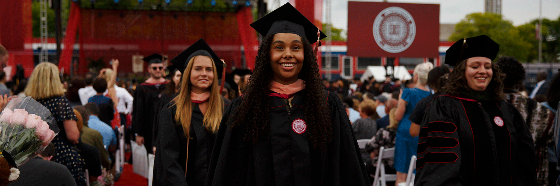 A female graduate and two others behind her smile as they walk down the aisle after the commencement ceremony.