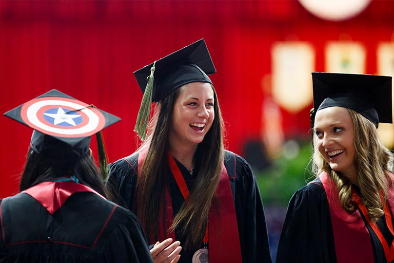 Three female undergraduate students smile at each other as their take their seats for Commencement