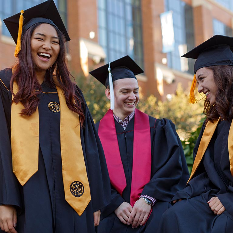 Three students in bachelor's commencement gowns gathered in a group outside  