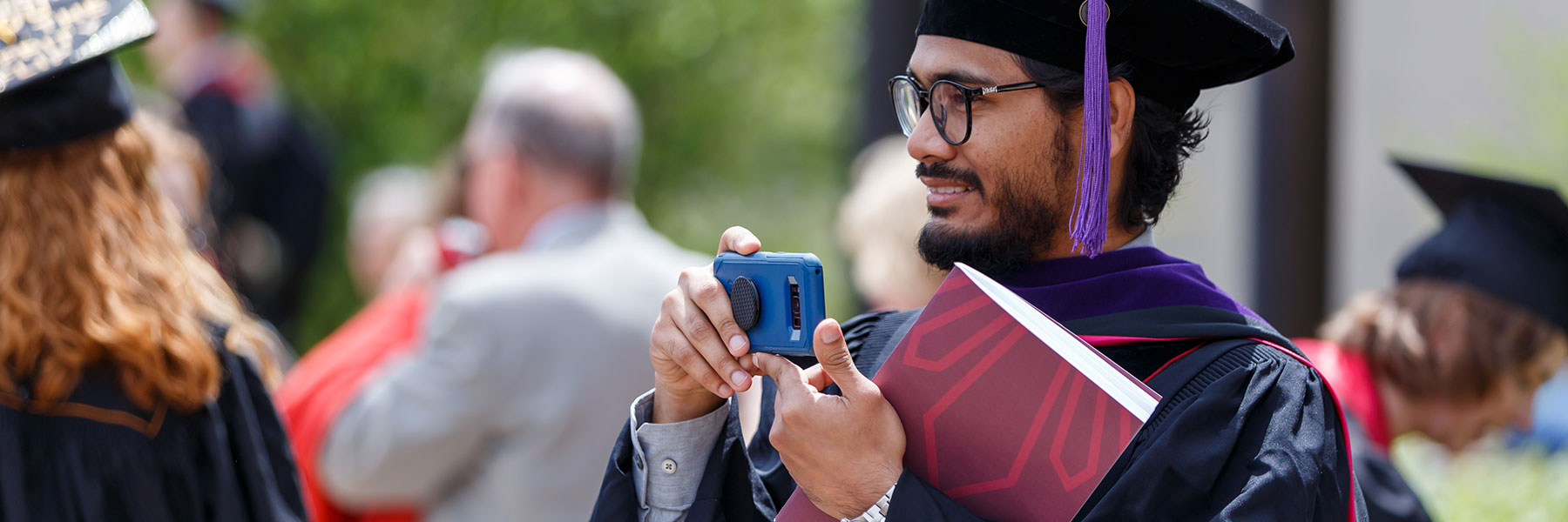 A student in commencement cap and gown holds a program booklet against his chest while holding up a phone to take a photo.