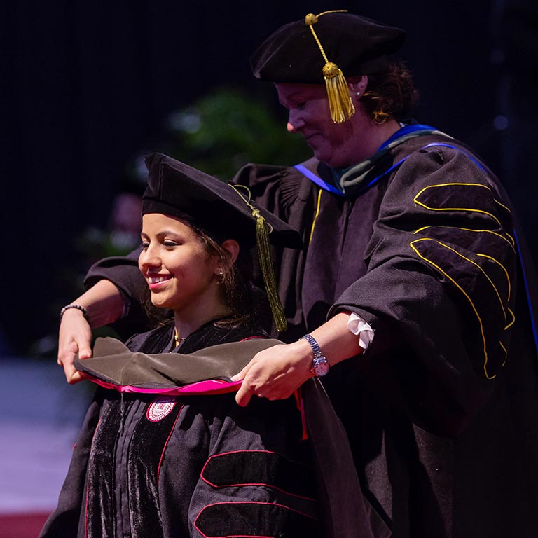 A woman in a doctoral robe looks forward as a faculty member behind her places a doctoral hood over her shoulders.