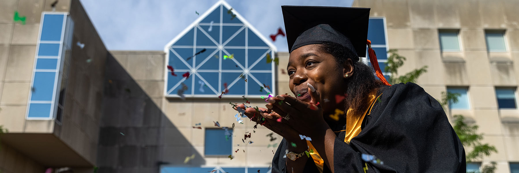 A graduate blows confetti from her cupped hands with the IUPUI University Library behind her.