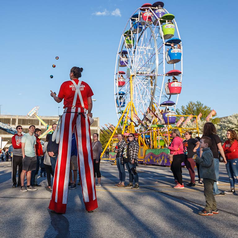 A performer on stilts juggles balls in the air; a ferris wheel is seen in the background.