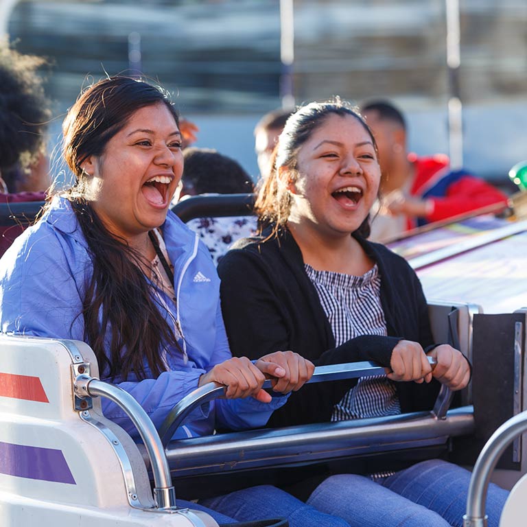 Two women sitting in a scrambler-style carnival game look forward with happy expressions.