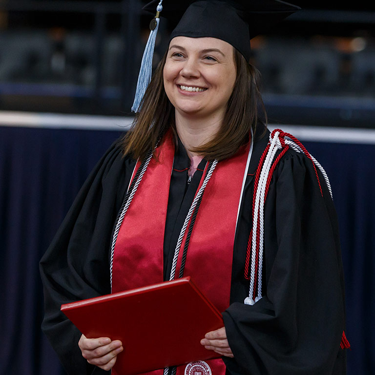 A woman holding a diploma cover in her hands looks ahead and smiles; she has red and white fourrageres on her shoulder
