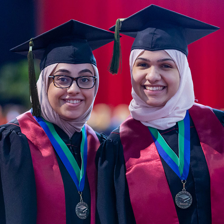 Two graduates look ahead smiling; both are wearing medallions hanging from blue and green ribbons around their necks.