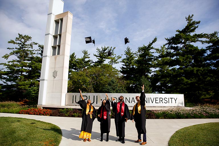 Four graduates thrown their caps in the air in front of the IUPUI gateway structure on the east side of campus.