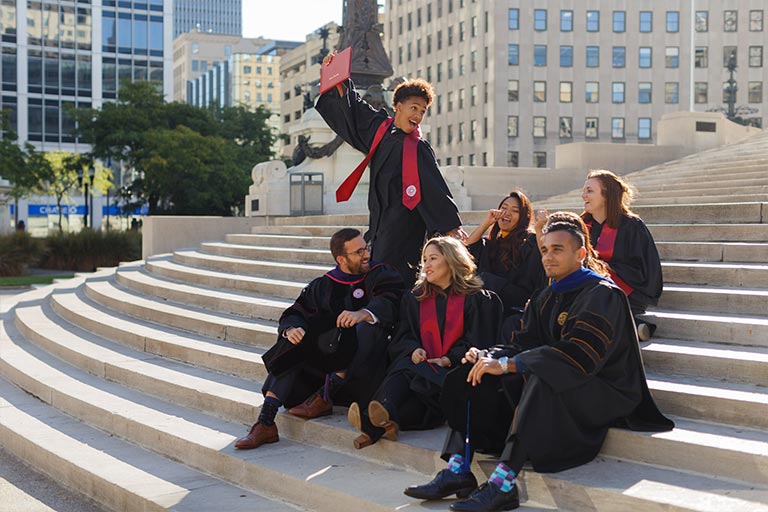 A group of graduates in commencement gowns sit on the steps of the Soldier's and Sailor's Monument in downtown Indianapolis; one student stands up holding his diploma