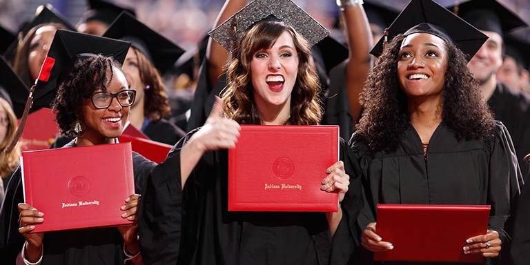 Three women in caps and gowns hold up crimson IU diploma covers smiling toward crowd