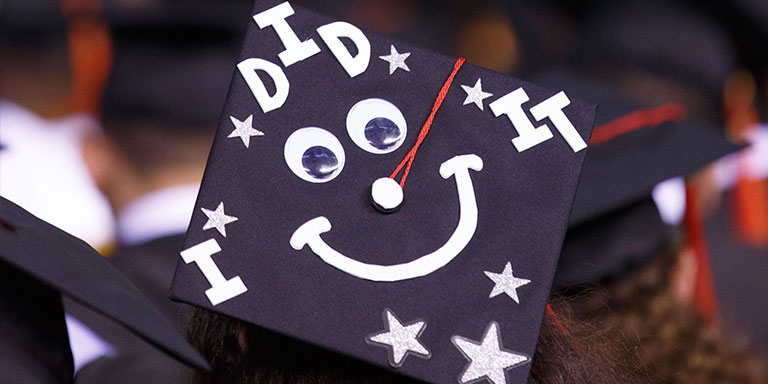 View of the back of a seated graduate's cap decorated with googly eyes and the words "I did it!"