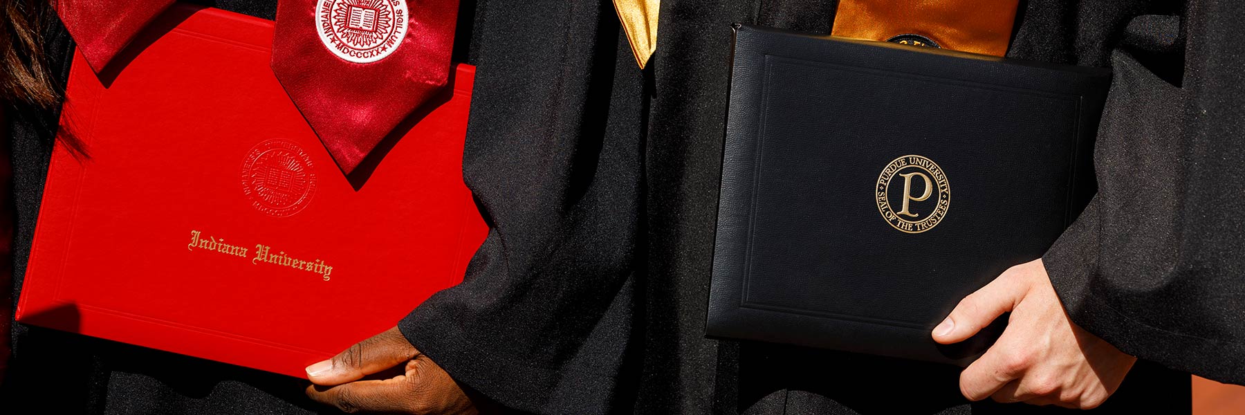 Detail image of two graduates holding diploma covers in their hand by their side—one diploma is for IU and one is for Purdue.
