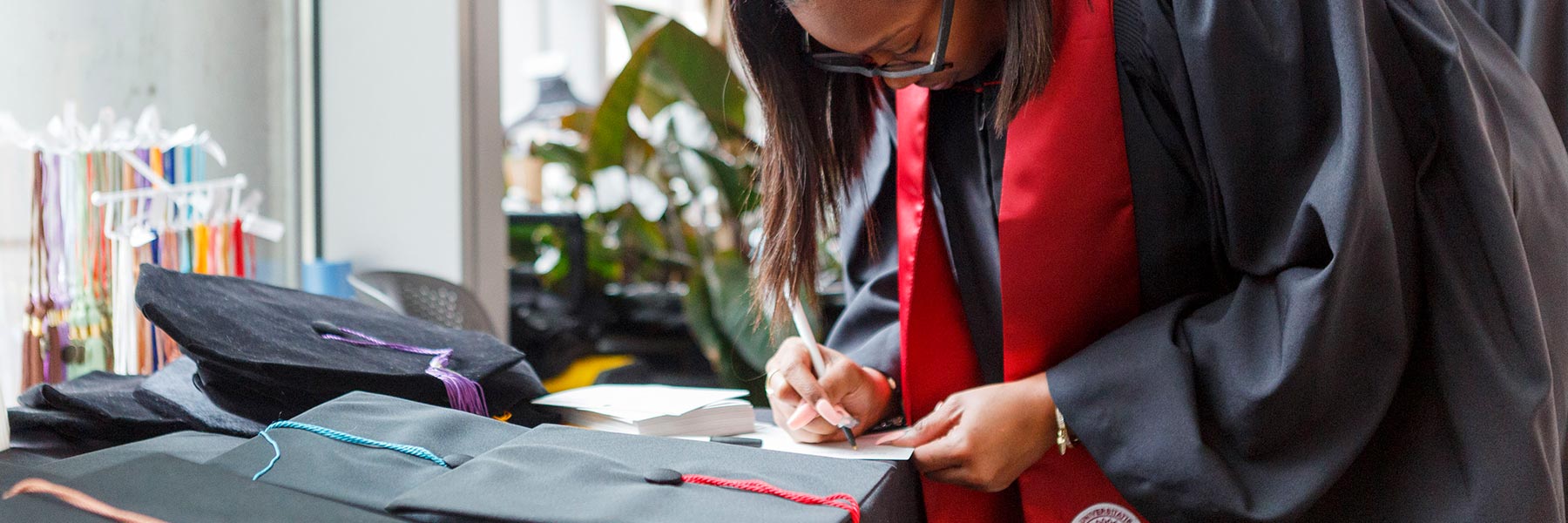 A graduate trying on a gown at GradFair leans down to write on a piece of paper at a table where tassels and caps are laid out.