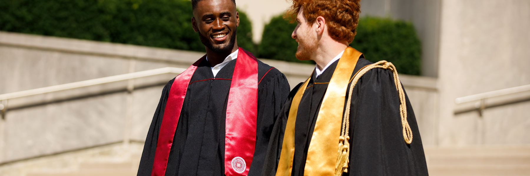 Two male graduates stand on outdoor steps; one is wearing an IU stole and the other is wearing a Purdue stole with honor cords.