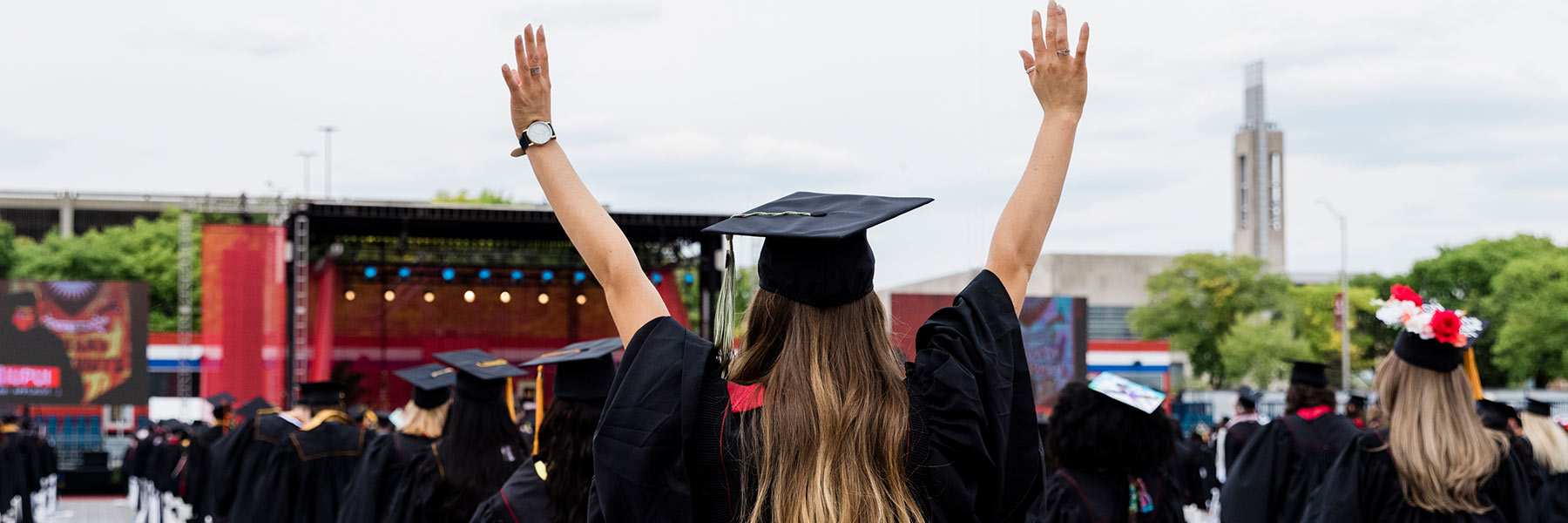 View of a graduate from behind holding her hands in the air as she stands looking at the commencement stage