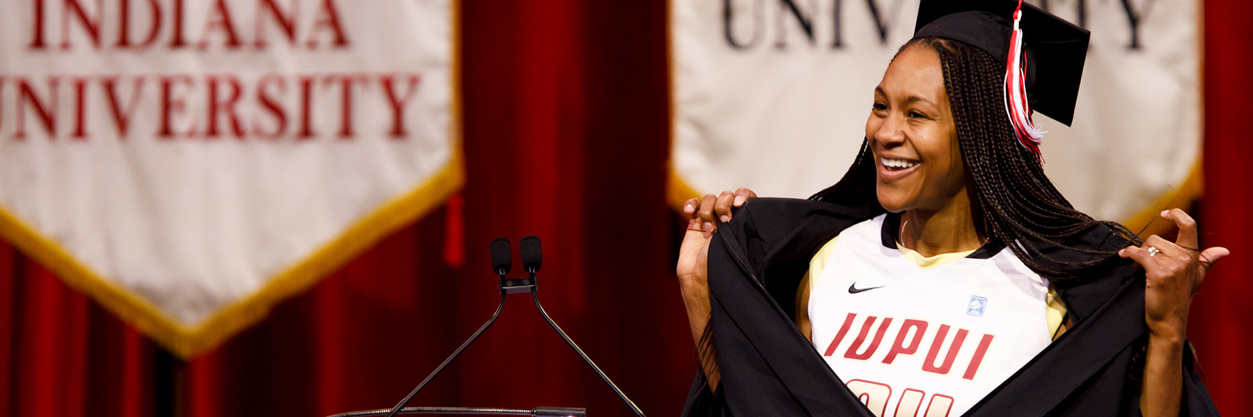 Past commencement speaker Tamika Catchings stands at a podium opening up her commencement gown to reveal an IUPUI Jags basketball jersey