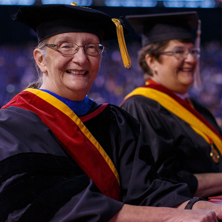 Two faculty in Commencement garb smile while seated for a ceremony